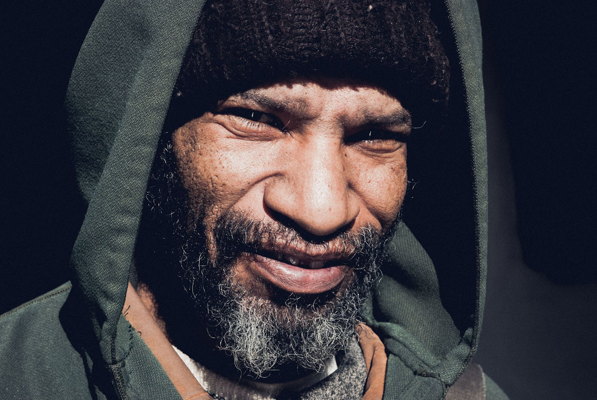 Close-up portrait of a man in a hooded jacket, weathered face lit against a dark background